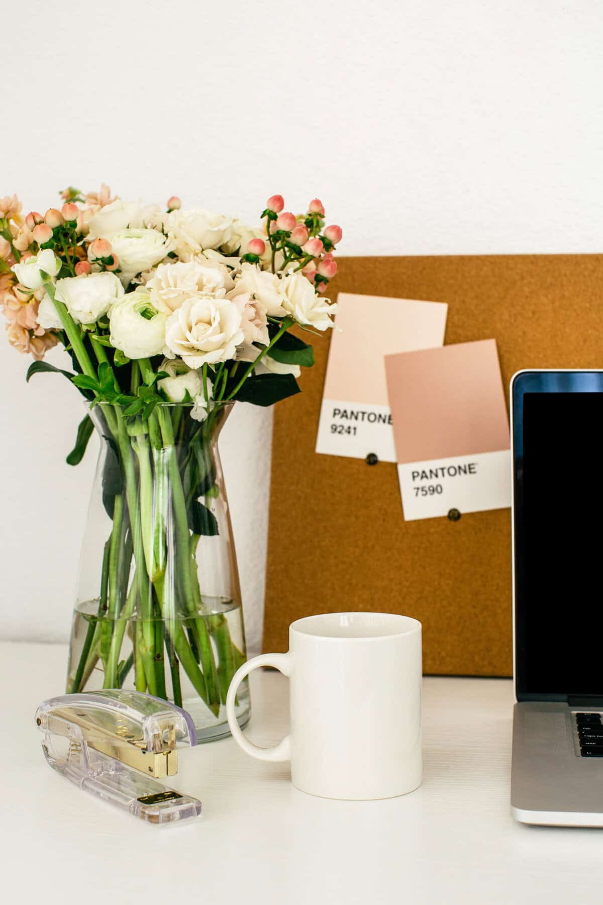 stock photo of part of a desk with a laptop, coffee mug, floral arrangement, and a cork board with pantone colors