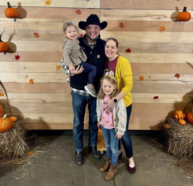 Photo of Melissa Mackey, her husband, and her two young daughters, standing in front of a wood backdrop